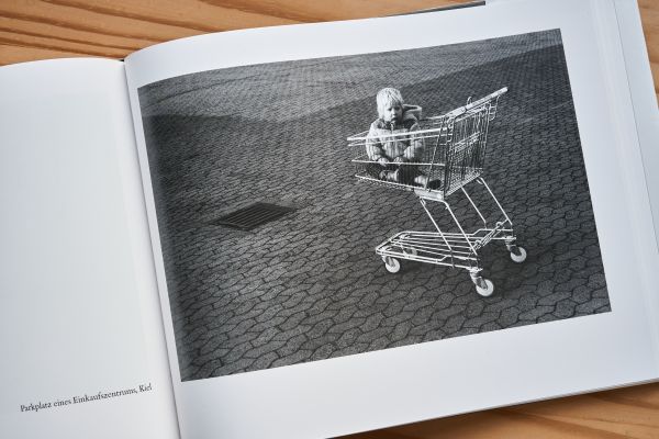 Ein kleines Kind sitzt allein in einem Einkaufswagen, umgeben von grauem Pflaster.